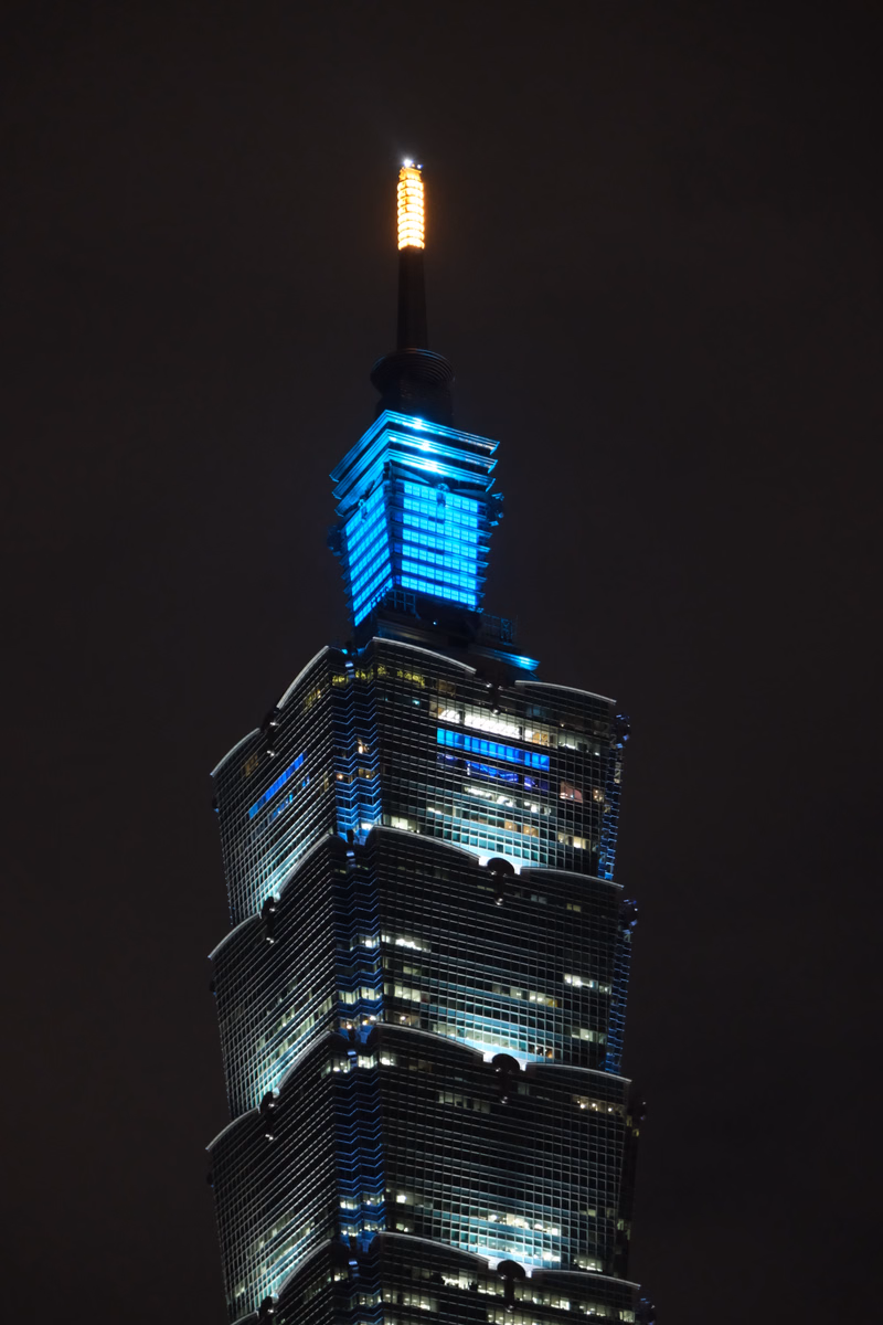 A modern skyscraper with illuminated windows and a bright spire stands against a dark night sky.