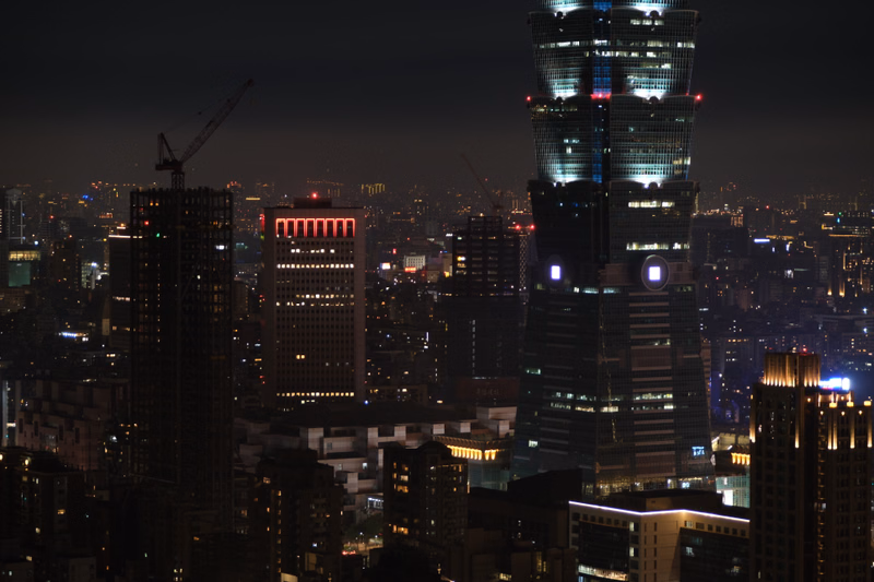 A tall, illuminated skyscraper dominates the night skyline of a dense city, surrounded by other buildings.