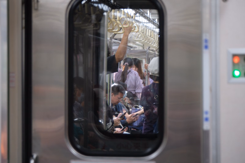A view through a train door shows several people inside a subway car, some looking at phones.