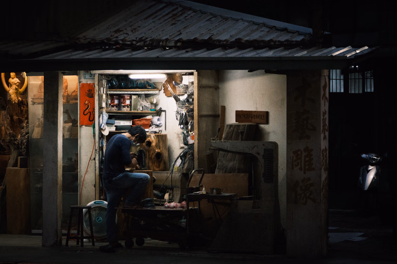 A craftsman works inside a dimly lit workshop, surrounded by wood and tools, under a corrugated metal roof.
