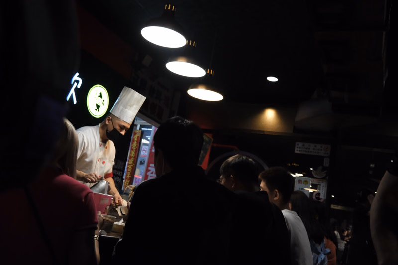 A chef wearing a tall white hat is working at a food stall under bright overhead lights, with a logo featuring a stylized character.
