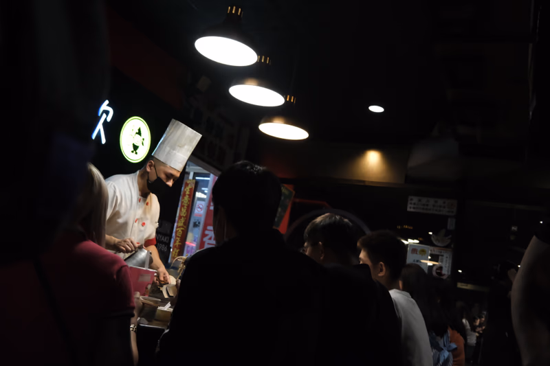 A dimly lit restaurant with a chef preparing food in the foreground and a group of people dining in the background.
