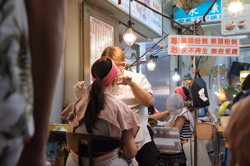 Two women stand outside a shop with a sign that reads "專員頭粉劑 黑頭粉劑 治永久再生 無效買費".