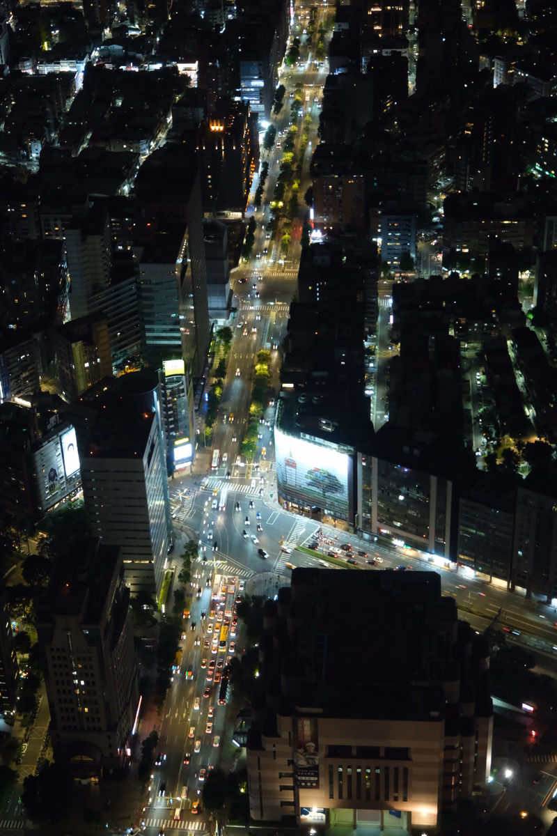 A high-angle nighttime view of a dense city street with numerous illuminated buildings and traffic.