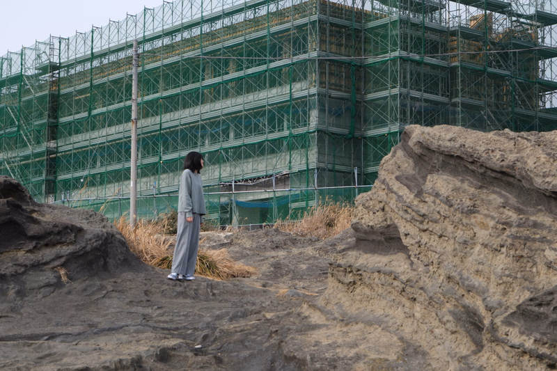 A woman in a grey outfit stands on sandy dunes overlooking a large building covered in green scaffolding.