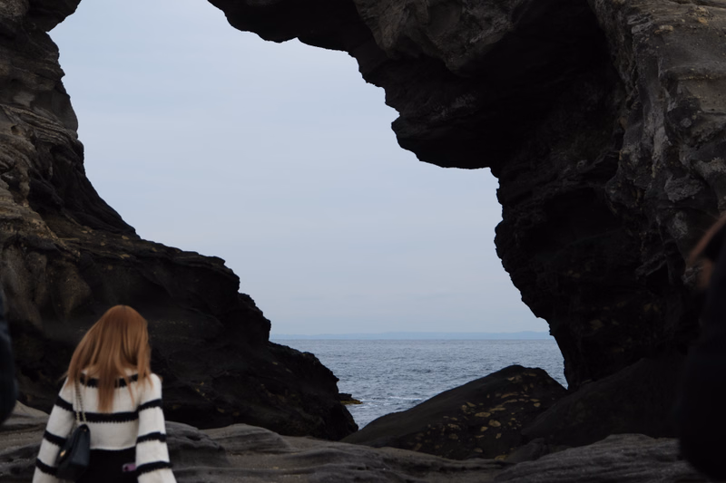 A woman in a striped sweater stands inside a large rock archway overlooking the ocean under a gray sky.