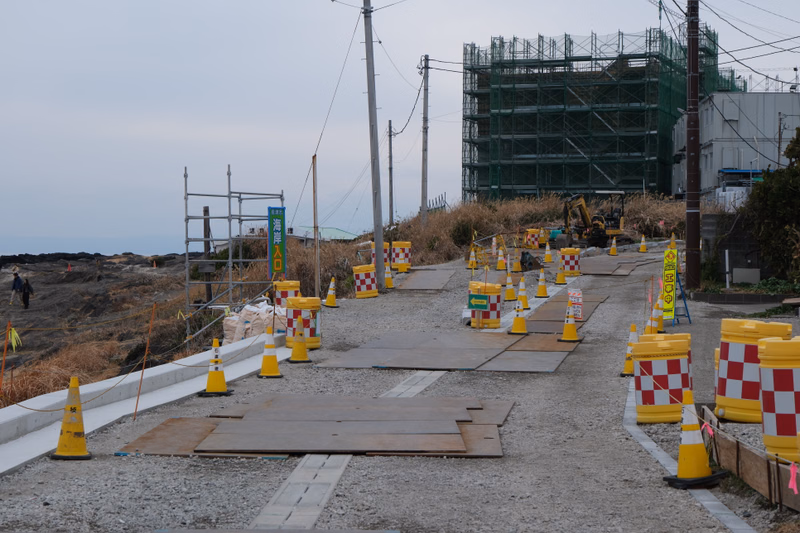 A construction site with scaffolding surrounding a large building, featuring a gravel road lined with yellow and red safety cones.