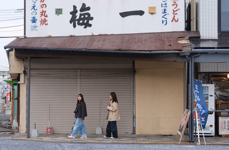 Three women stand outside a restaurant with a sign that reads "梅 一" and "の丸焼".