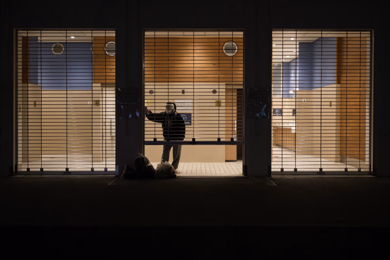 A photograph of a person standing in a room with blinds, looking out the window.
