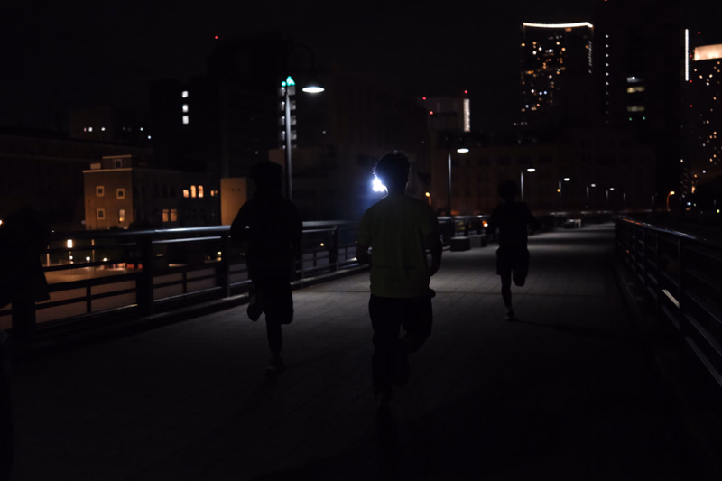 Three runners are jogging along a paved path at night with city lights visible in the background.