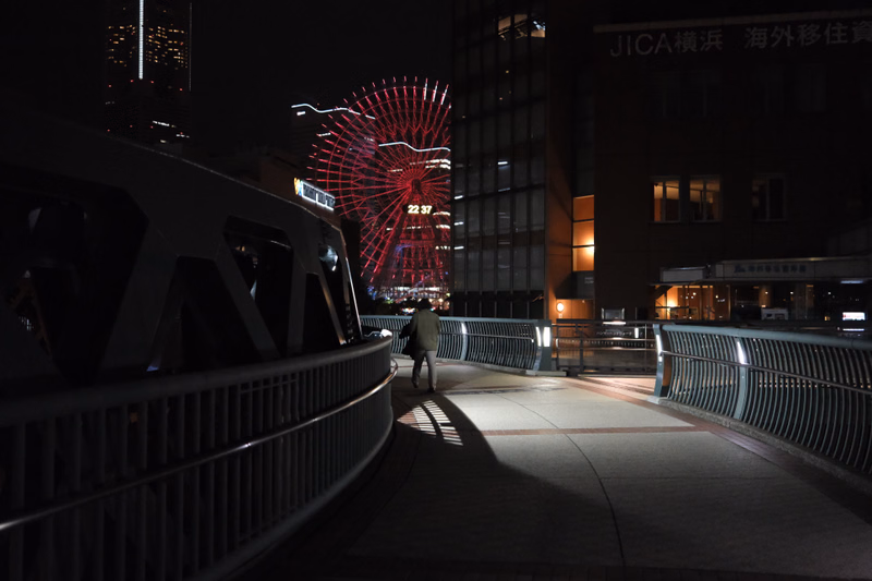 A person walks along a curved bridge at night, with a large illuminated red display visible in the background.