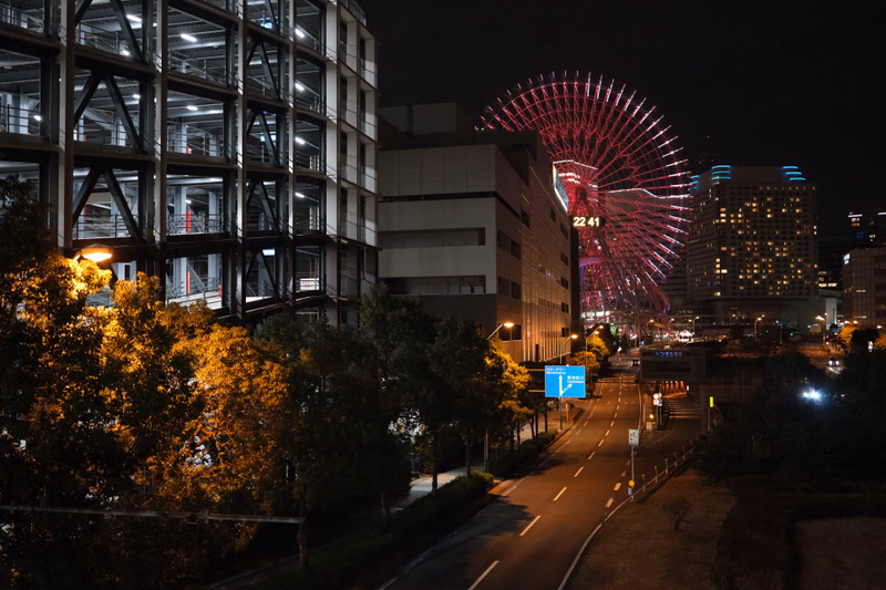A large Ferris wheel with red lights illuminates a city street at night, next to modern buildings.