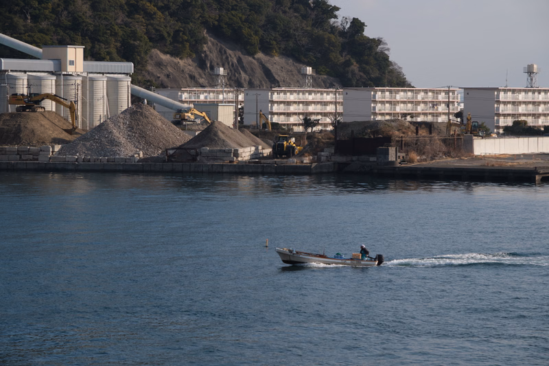 A small motorized boat travels across the water near an industrial area with large storage tanks and buildings.