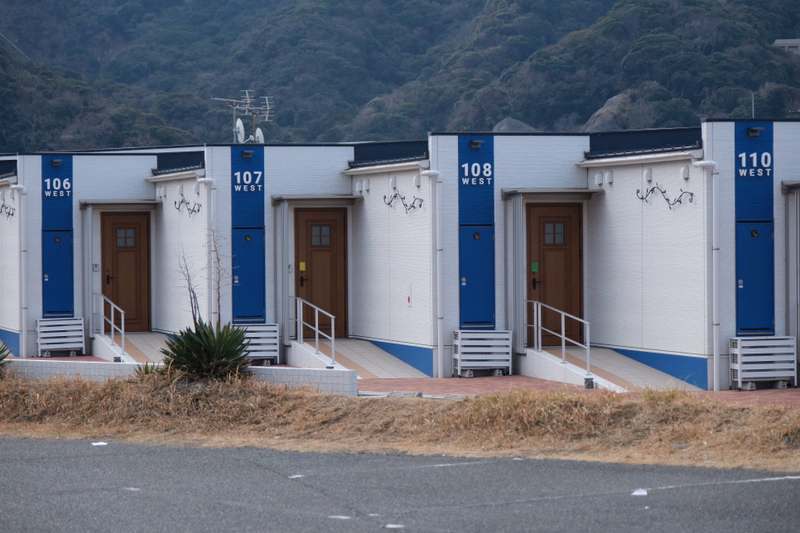 A row of modern beach huts, numbered 106 through 110, are situated in front of a steep, green hillside.