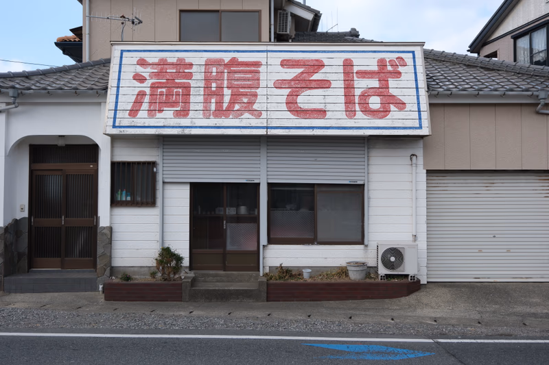 A small building with a sign that reads 'Kurihama' in Japanese. The building is located near a road.