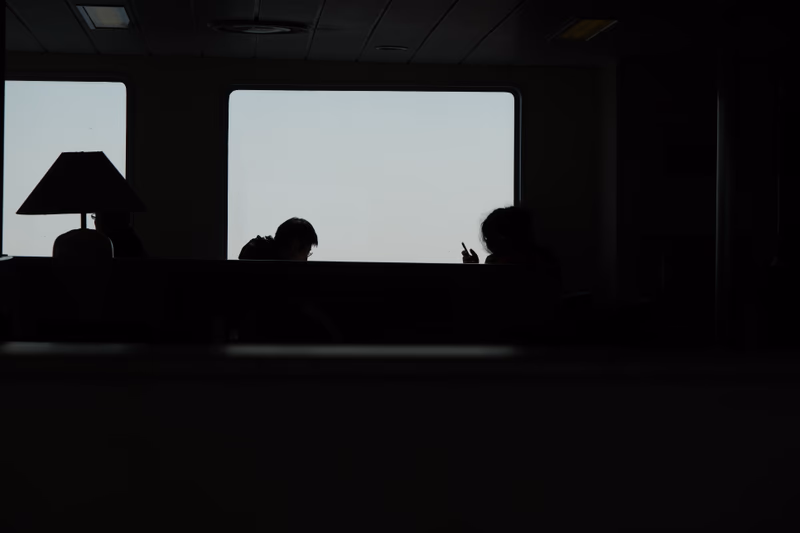 A silhouette of people sitting by a large window with a lamp on the left side.