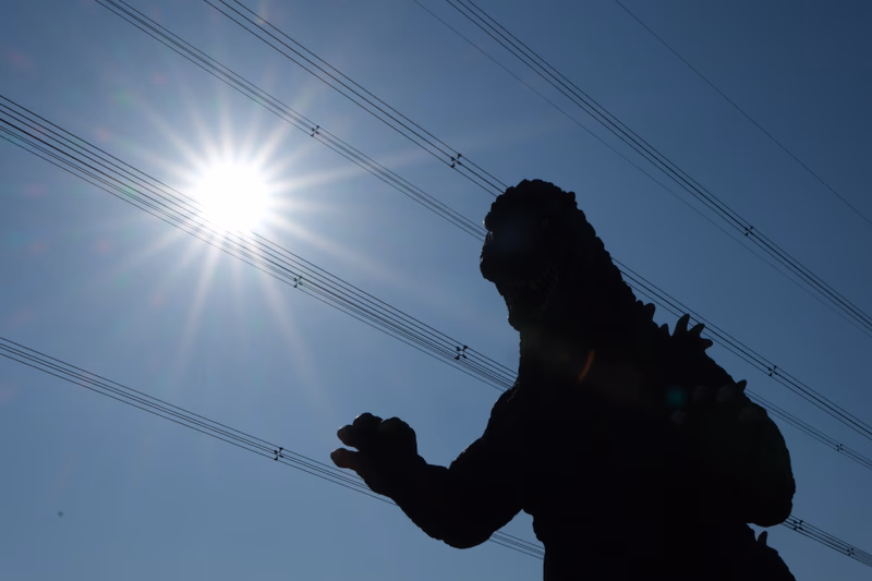 A statue of a creature resembling Godzilla stands near power lines on a sunny day.