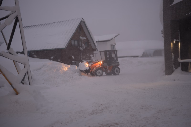A small orange tractor is parked in the snow near a dark wooden house during a heavy snowfall.