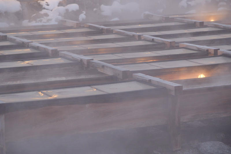 A wooden outdoor sauna structure is enveloped in thick steam, with warm light visible from the interior.