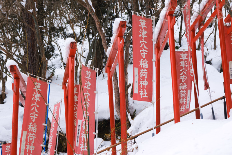 Several red torii gates stand in a snowy outdoor setting, with signs reading "草津八宇稲荷神社".