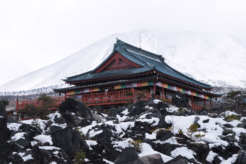 A traditional Japanese temple with colorful eaves is situated on a rocky slope against a backdrop of a snow-covered mountain.
