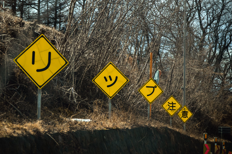 A series of yellow diamond-shaped Japanese warning signs are posted along a roadside, featuring characters like "リン", "ツッ", "プ", and "注意".