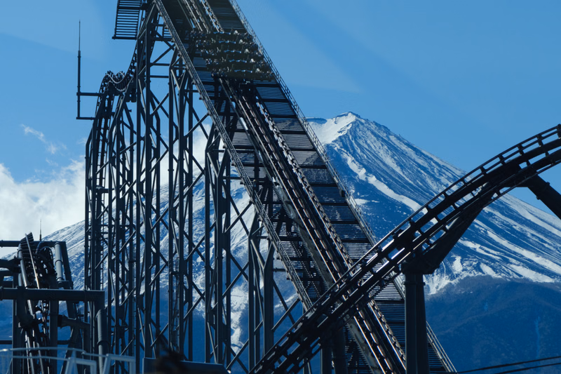 A large steel roller coaster climbs steeply against a backdrop of a snow-capped mountain under a clear blue sky.