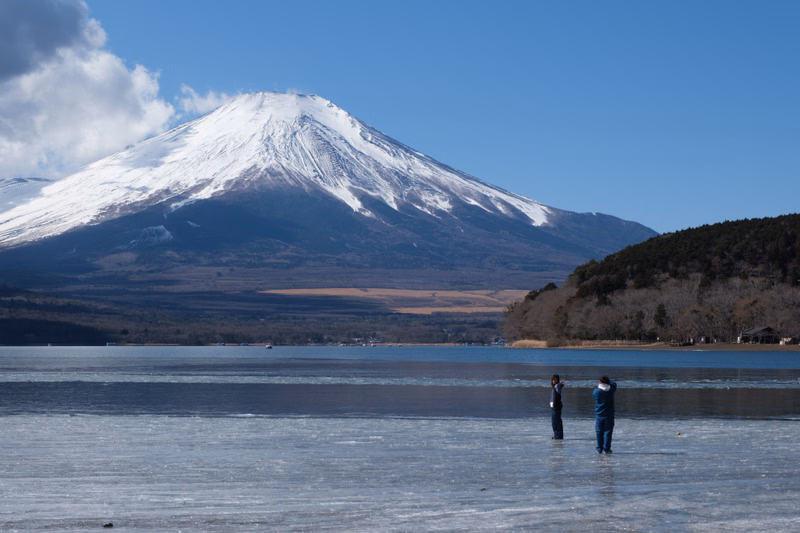 Two people stand on a frozen lake in the foreground with a large, snow-capped volcano dominating the background under a clear blue sky.