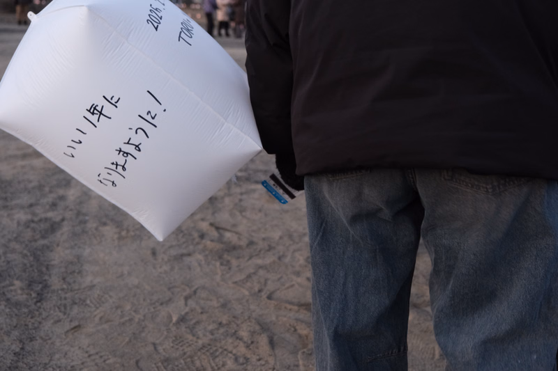 A white balloon with Japanese text reading "いい年をありがとう!" is held near a person standing on the sand.