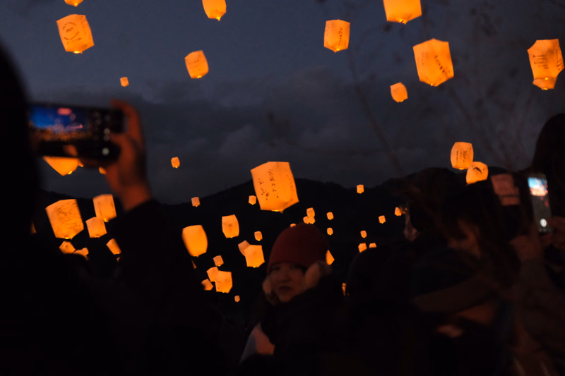 Many glowing sky lanterns float in the dark night sky above a crowd of people attending a festival.