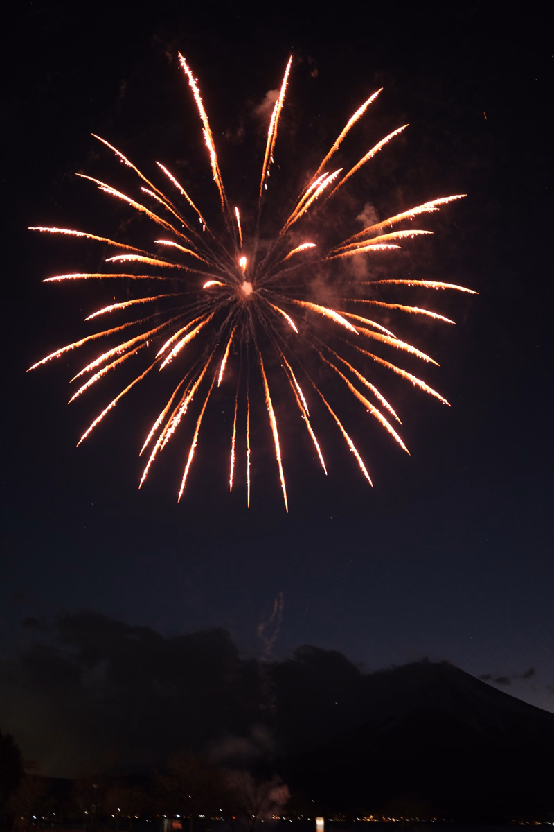 A large, bright burst of fireworks illuminates the dark night sky above a silhouette of mountains.