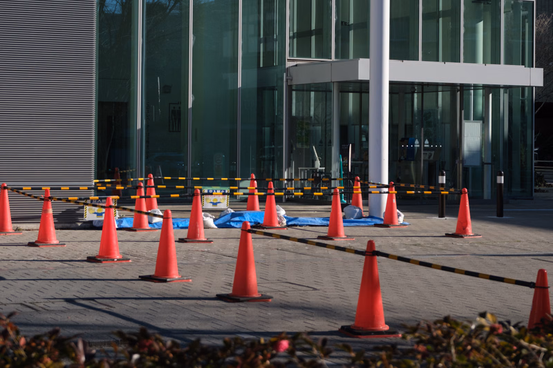 Several orange traffic cones are placed on the paved area in front of a modern glass facade building entrance.