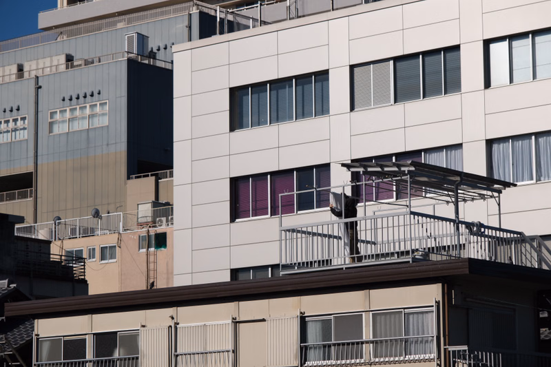 A modern, white apartment building features multiple balconies and rows of windows, with another large building visible in the background.