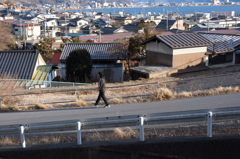 A person walks along a road overlooking a dense cityscape and a body of water with many houses.