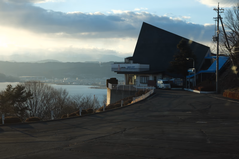 A photo of a road leading to a building with a unique, angular design, overlooking a lake and mountains.