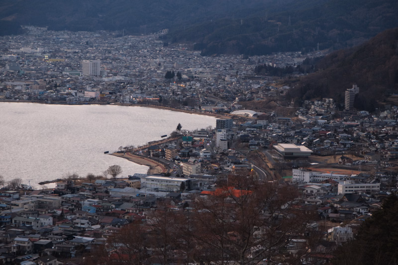 A wide view of a densely packed town situated along the shore of a large body of water, with hills rising in the background.