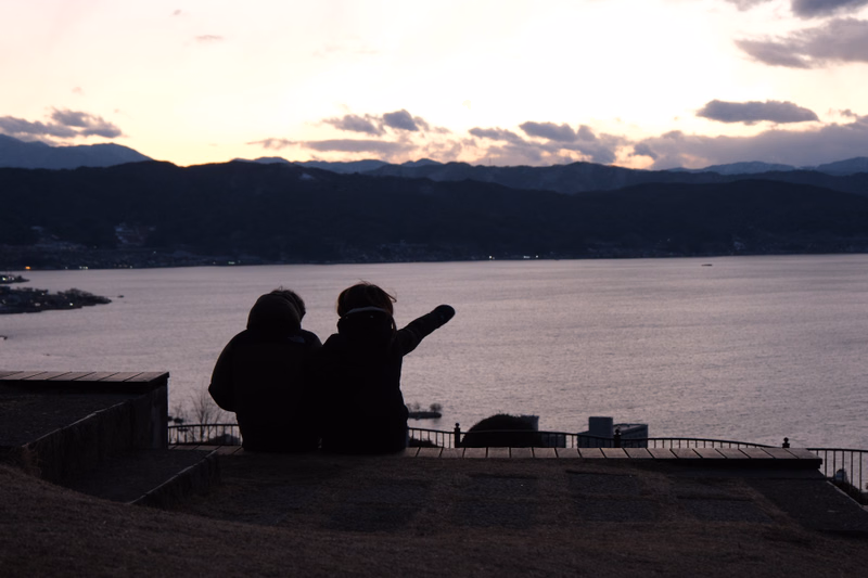 Two people stand on a grassy hill overlooking a large body of water during sunset, with mountains in the background.