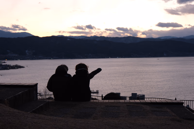 Two people sitting on a hill overlooking a calm sea with mountains in the background.