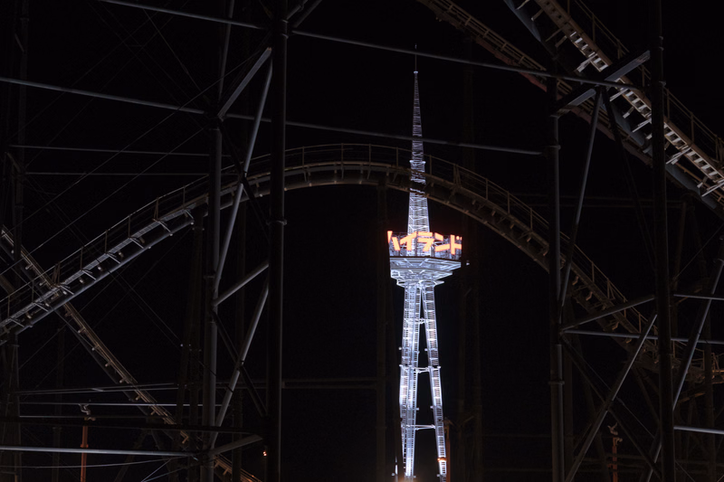 A tall, illuminated tower stands against the dark night sky, framed by the dark metal structure of a bridge.