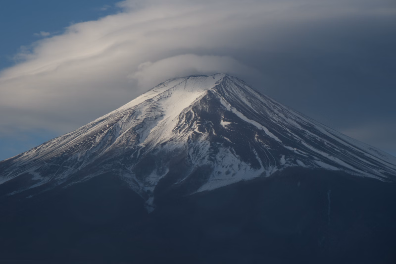 A snow-capped mountain, likely a volcano, is partially shrouded in low-hanging clouds under a blue sky.