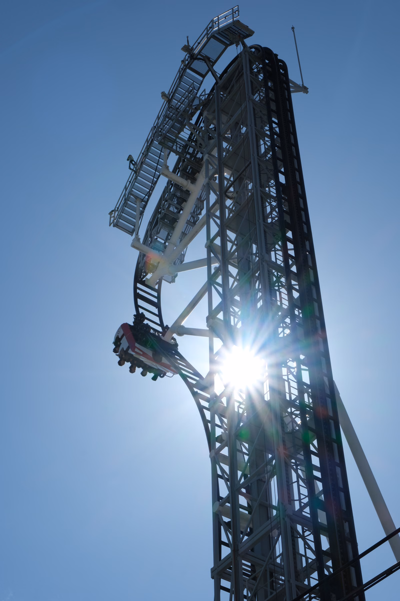 A large steel roller coaster structure dominates the frame against a bright blue sky, with the sun flaring through the supports.