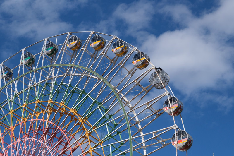 A large Ferris wheel and a roller coaster are visible against a blue sky with white clouds at an amusement park.