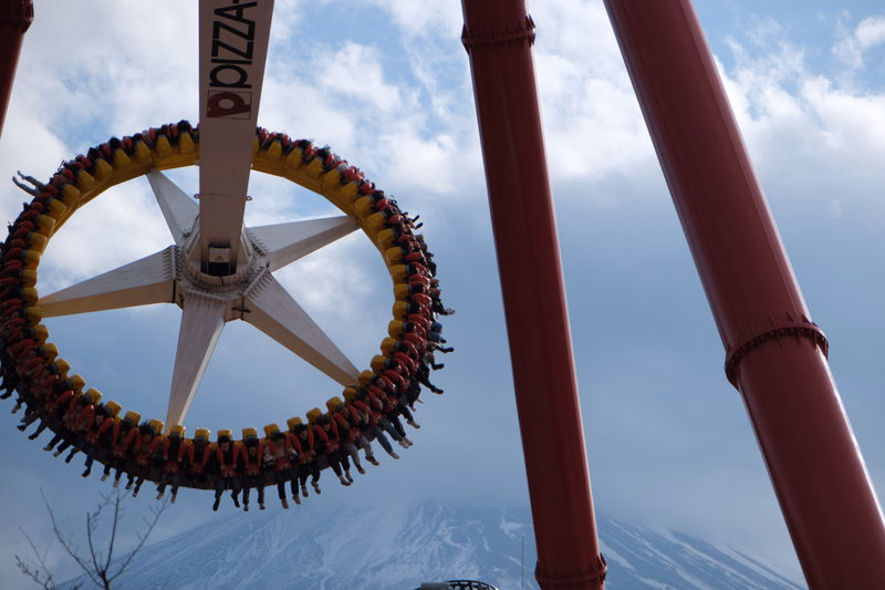 A large pizza-themed wheel sign, featuring the text "PIZZA", hangs in front of tall red roller coaster supports against a cloudy sky.