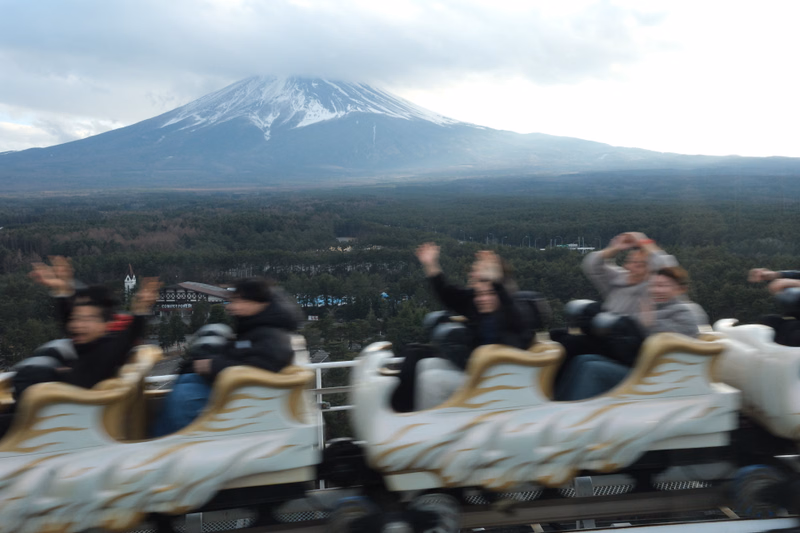 A panoramic view of a snow-capped mountain, likely Mount Fuji, seen from a tourist train overlooking a forested landscape.
