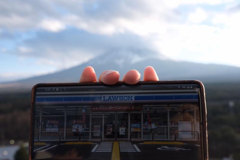A smartphone displaying the Lawson convenience store logo is held against a backdrop of a large, hazy mountain under a cloudy sky.