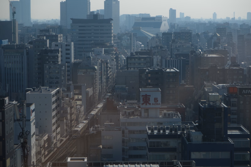 A dense cityscape view of numerous high-rise buildings under a hazy sky, with a sign visible that reads "東 5 AZMA".