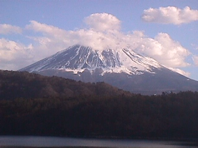 A snow-capped mountain, likely a volcano, rises above a dark forest bordering a calm lake under a partly cloudy sky.