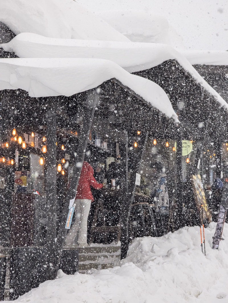 A building with snow-covered eaves is visible during a heavy snowfall, with a person standing near the entrance.