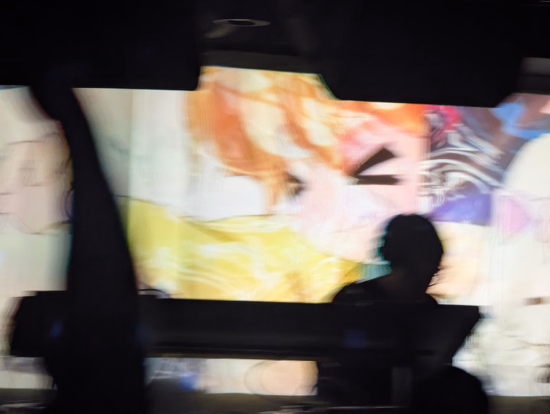 A brightly colored, blurred illustration of an anime-style person with orange hair, viewed through a dark foreground obstruction.