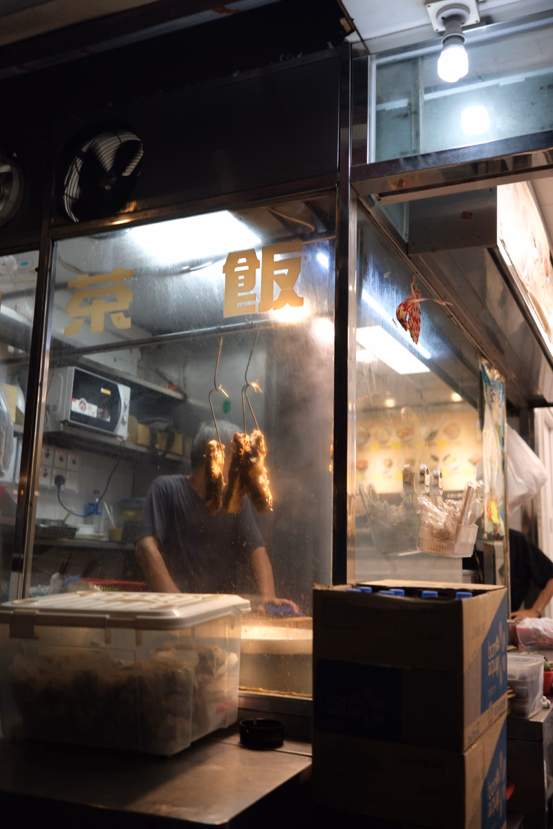 A person is frying fish inside a glass shop with Chinese characters visible on the window, reading "飯".